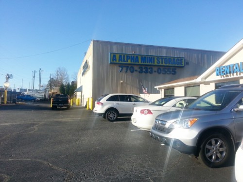 Outdoor signage for 'Alpha Mini Storage' by Pinnacle Custom Signs, prominently displayed on the front of an industrial building. The sign features bold, blue letters with a phone number, visible over a busy parking lot filled with cars and a clear blue sky in the background.